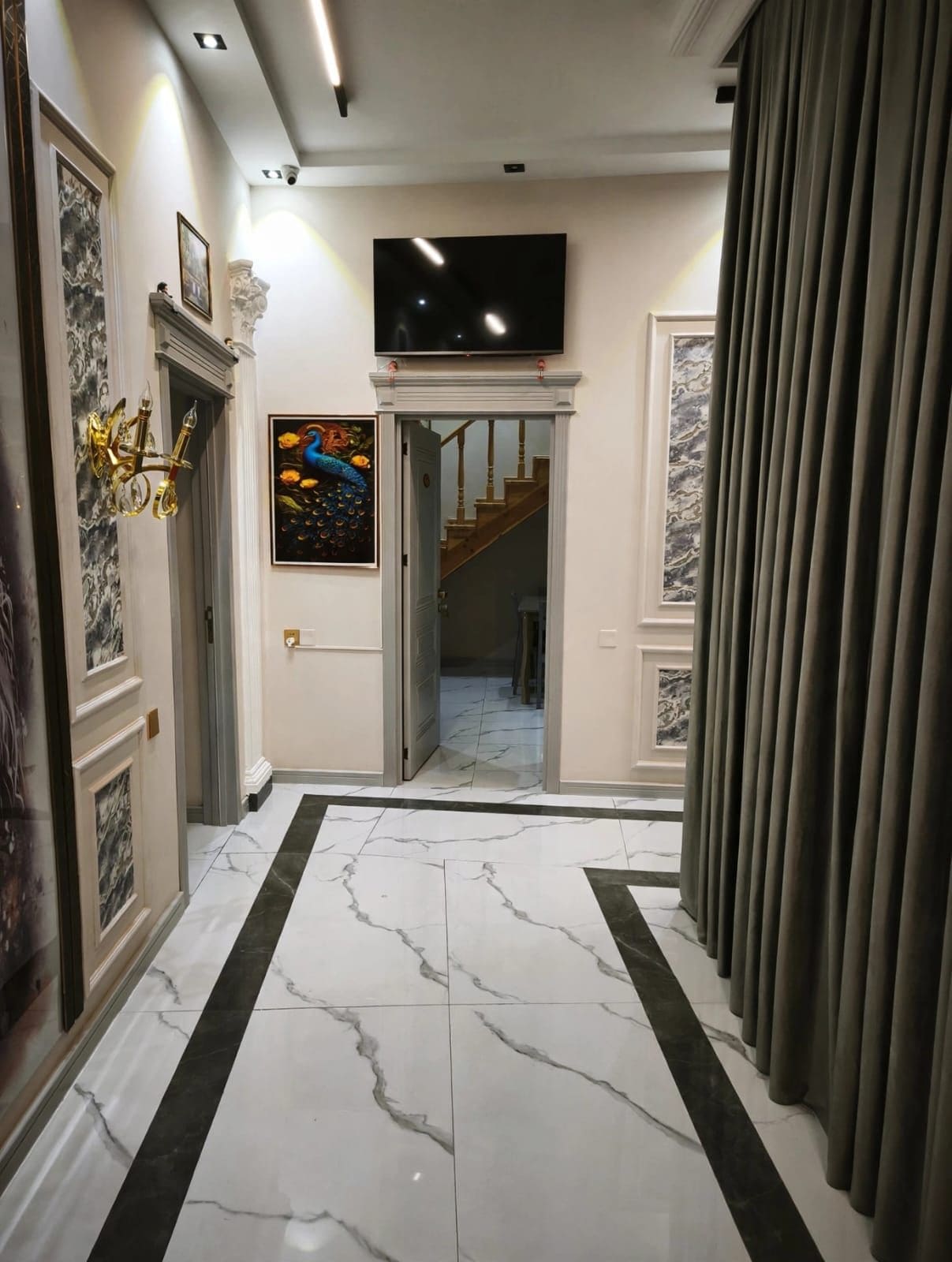 Um corredor com piso de mármore, uma TV de tela plana instalada acima de uma porta e uma escada que leva a outro cômodo.