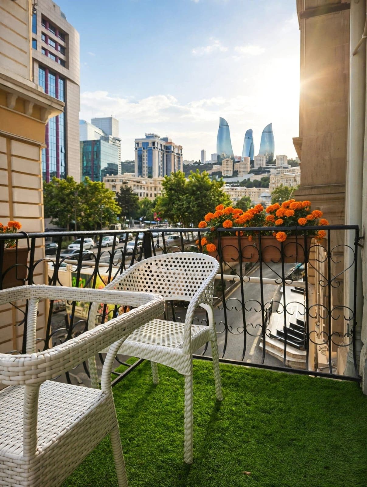 Un balcón con sillas de mimbre blanco, una jardinera con flores naranjas y vistas a la ciudad que incluyen edificios modernos y las Torres Llama de Bakú.