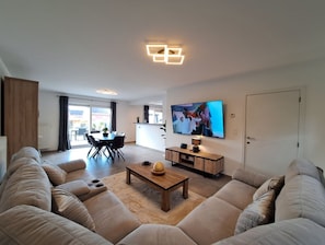 Una sala moderna con un sofá modular, una mesa de centro, un televisor de pantalla plana montado en la pared y un área de comedor al fondo.