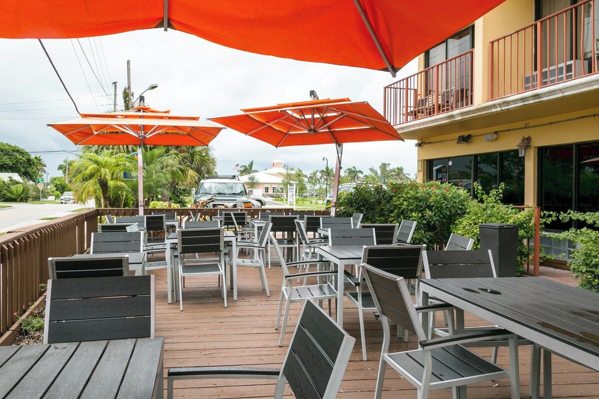 An outdoor patio with tables and chairs, covered by orange umbrellas, and a building with a balcony in the background.