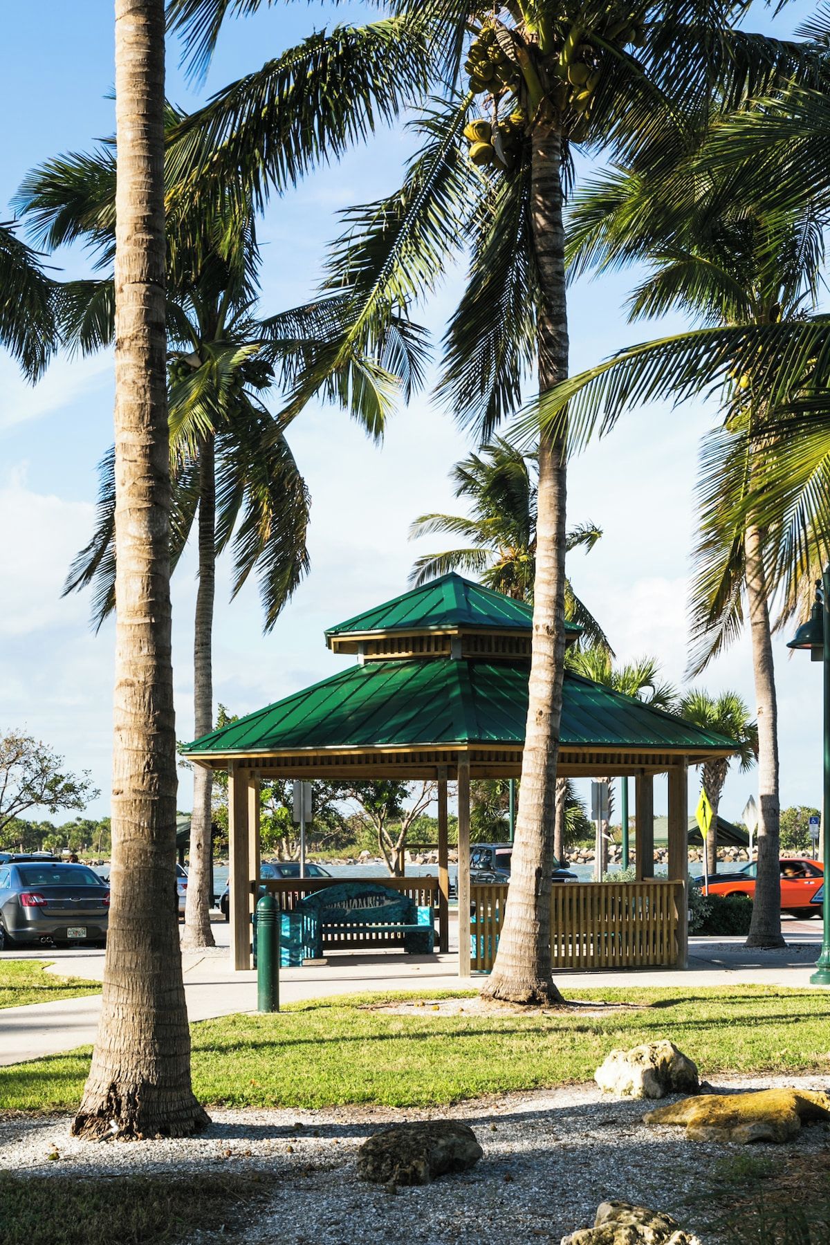 A gazebo with a green roof, surrounded by palm trees and a gravel path.
