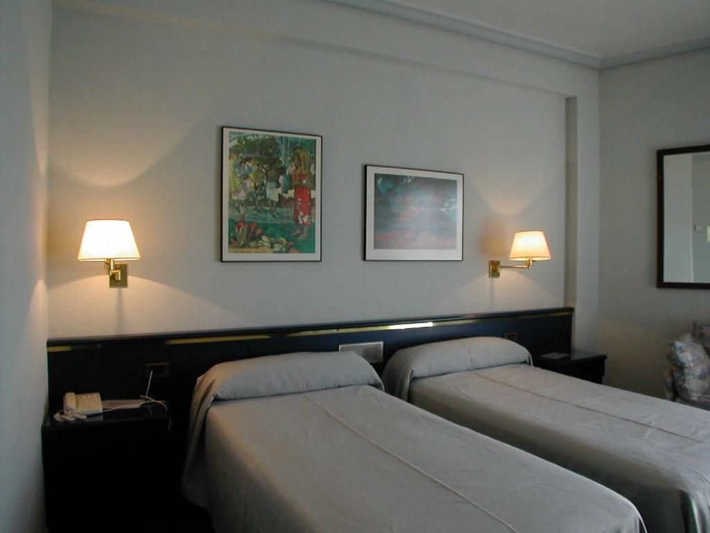 A hotel room with two single beds, a nightstand with a lamp, and two framed pictures on the wall.