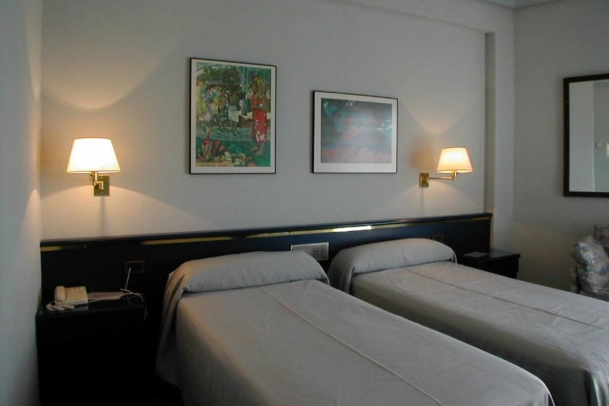 Quarto de hotel com duas camas de solteiro, mesa de cabeceira com abajur e dois quadros na parede.