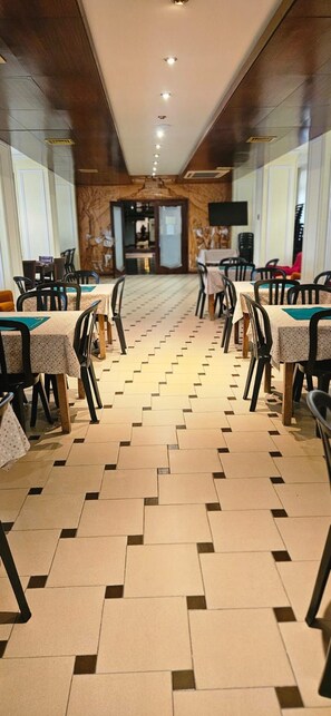 Corredor de hotel com mesas e cadeiras dispostas para refeições, piso estampado e parede de madeira com uma porta no final.