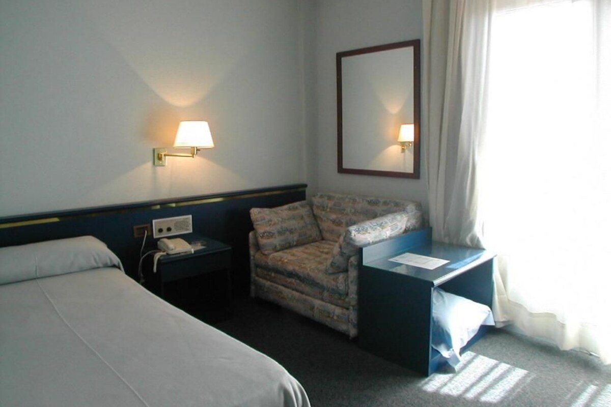 Quarto de hotel com cama, cadeira, escrivaninha, luminária, telefone e espelho.