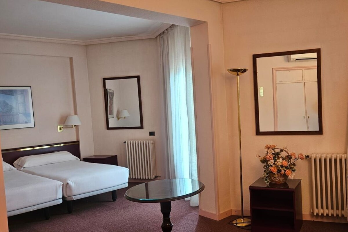 Quarto de hotel com cama, escrivaninha com televisão, mesinha, espelho e radiador.