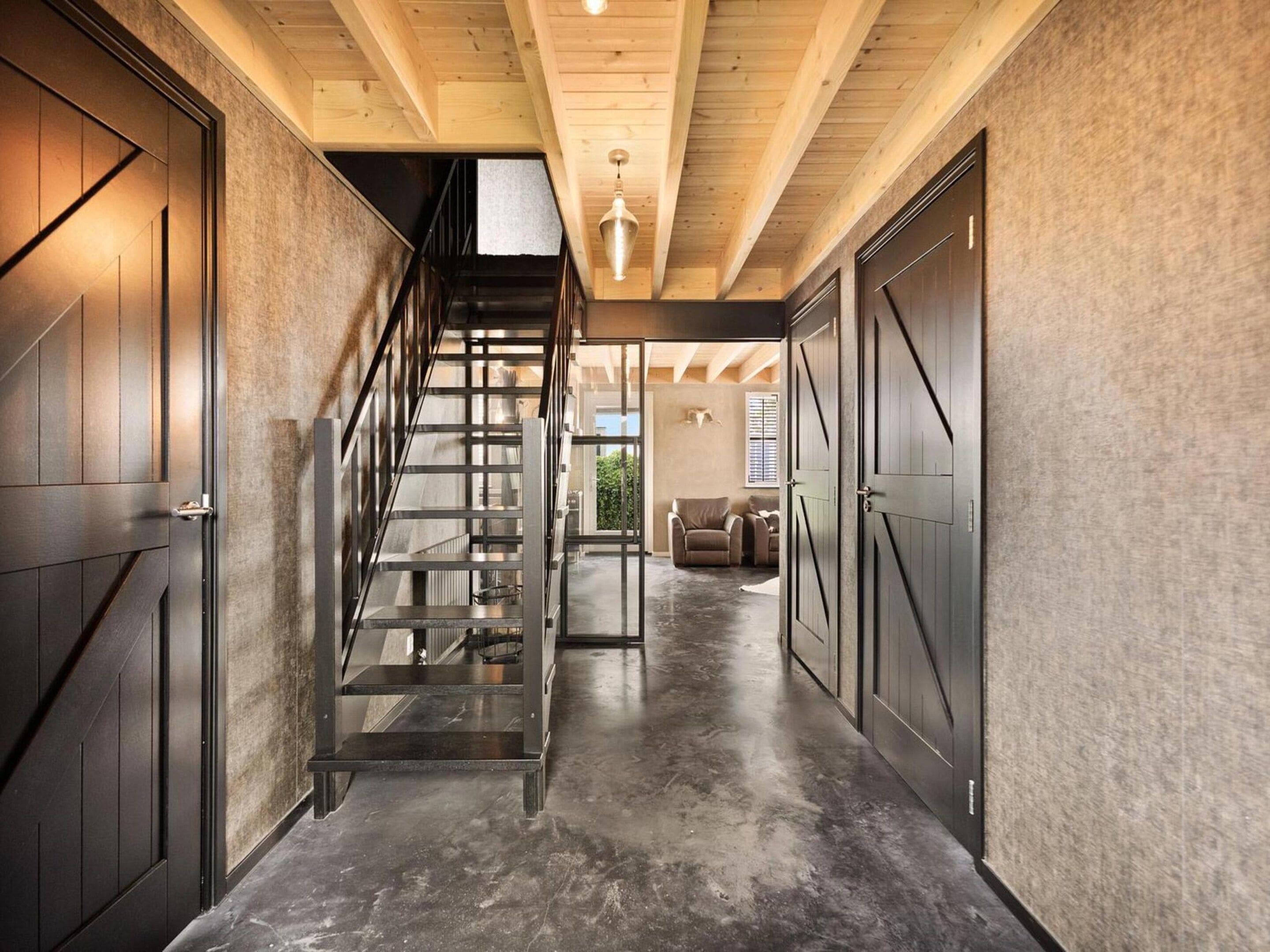 Un intérieur moderne avec un escalier menant à une mezzanine, un coin salon avec un canapé et des fauteuils, et une grande porte sur le côté droit.