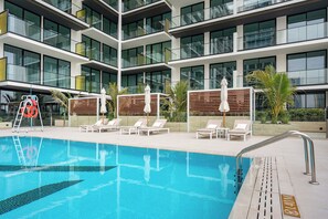 A modern hotel pool area with multiple lounge chairs and umbrellas, surrounded by a multi-story building with large windows. - Designer 1BR, Pool View, Finest Renovation JVC (Dubai)