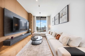 A modern living room with a large flat-screen TV, a sofa, a coffee table, and a view of the city through large windows. - Designer 1BR, Pool View, Finest Renovation JVC (Dubai)