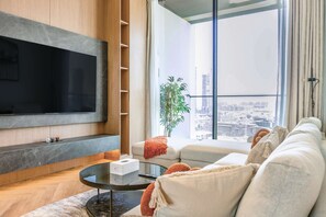 A modern living room with a flat-screen TV mounted on a wall, a glass coffee table, a sofa with cushions, and a potted plant. - Designer 1BR, Pool View, Finest Renovation JVC (Dubai)