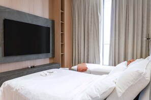 Camera d'albergo con una grande TV a schermo piatto fissata al muro, due letti con lenzuola bianche, una parete rivestita in legno e una finestra con tende.