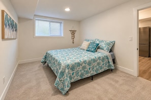 Un dormitorio con cama, ventana, lámpara de pared y puerta que da a una cocina.