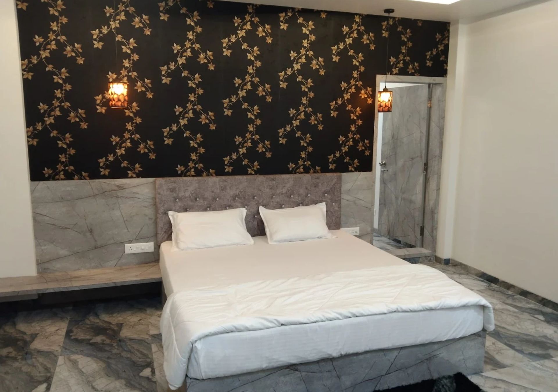 Un dormitorio con una cama, dos almohadas, suelo de mármol, pared de un diseño oscuro y dos luces fijadas a la pared.