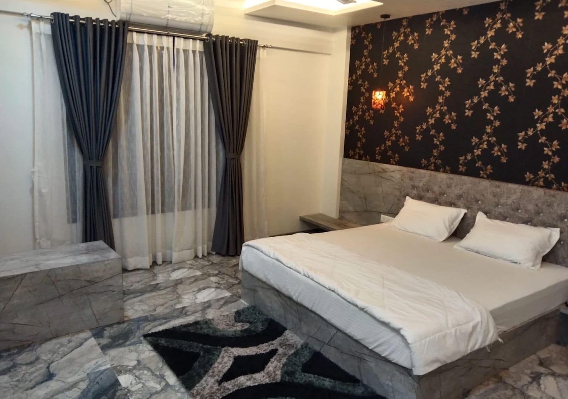A hotel room with a bed, curtains, a patterned wall, and a nightstand.