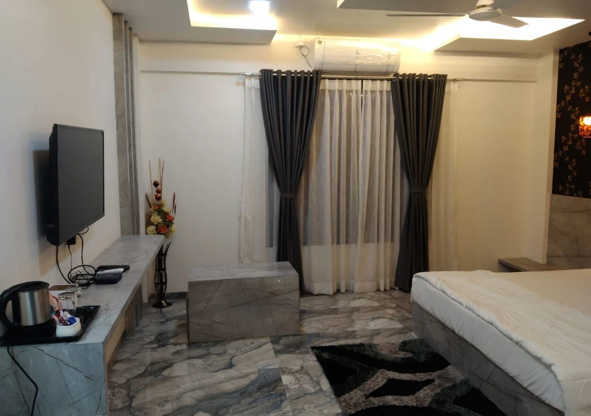 Une chambre d’hôtel équipée d’un lit, d’un téléviseur à écran plat, d’un sol carrelé en marbre et d’une grande fenêtre avec des rideaux.