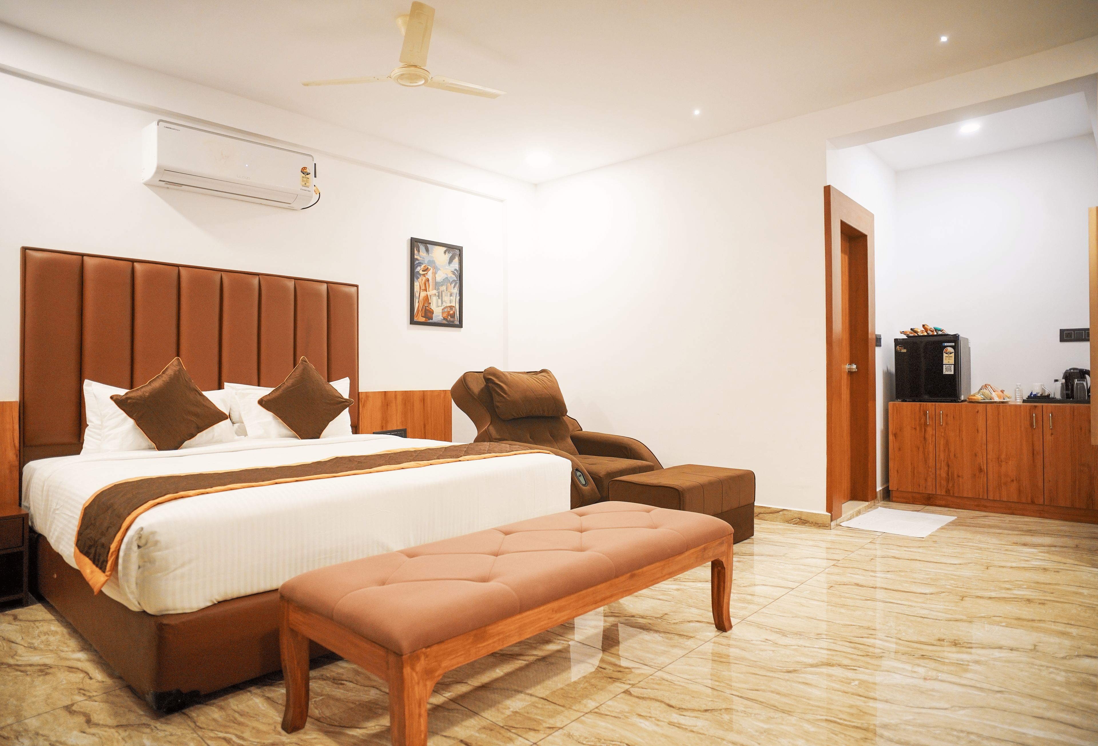 A hotel room with a large bed, a bench, a chair, a ceiling fan, and a painting on the wall.