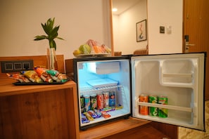 A compact refrigerator with a variety of snacks and beverages inside, a small plant on top, and a wooden counter beside it. - Bliss Hotel (Bengaluru)