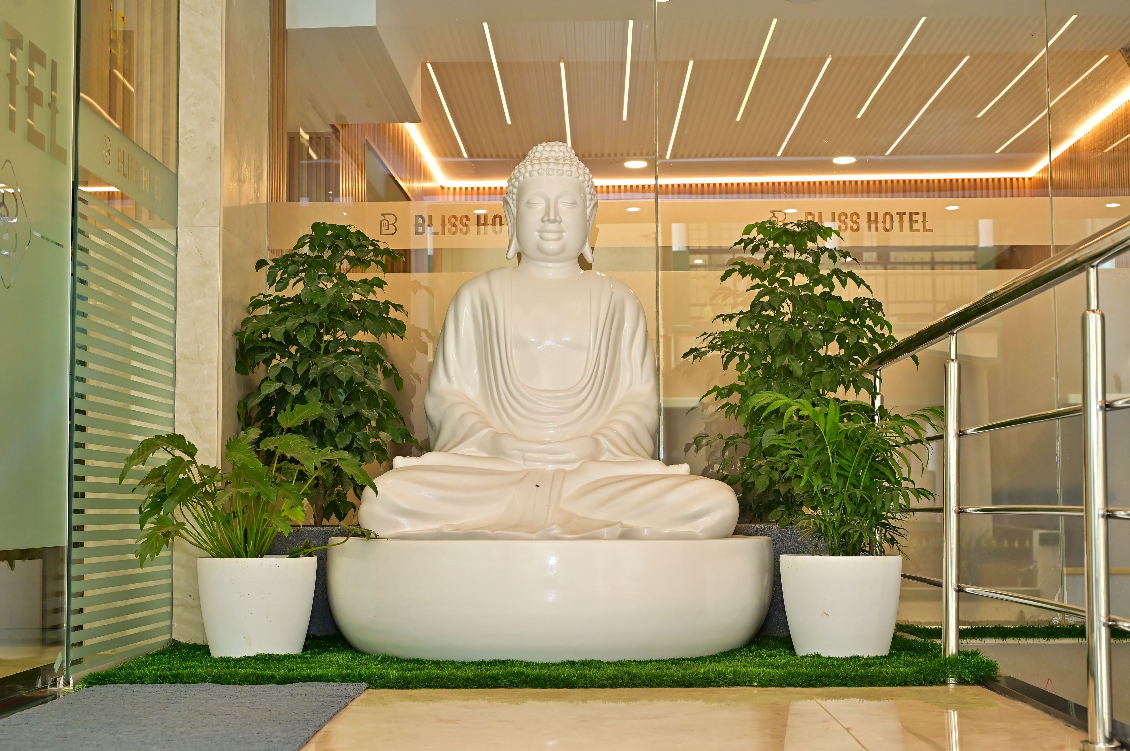 A Buddha statue in a modern lobby with a glass wall, potted plants, and a staircase.