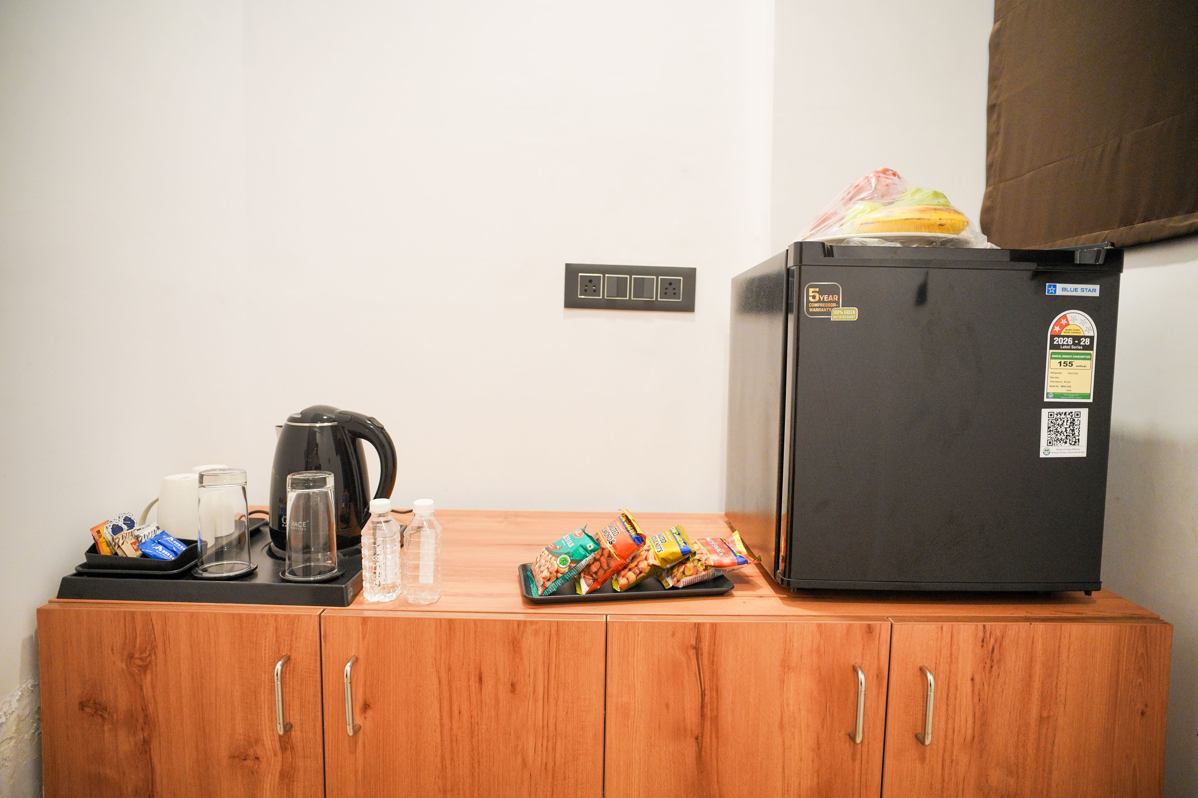 Uma cozinha compacta com geladeira preta, cafeteira, chaleira e embalagens de lanches em uma bancada de madeira.