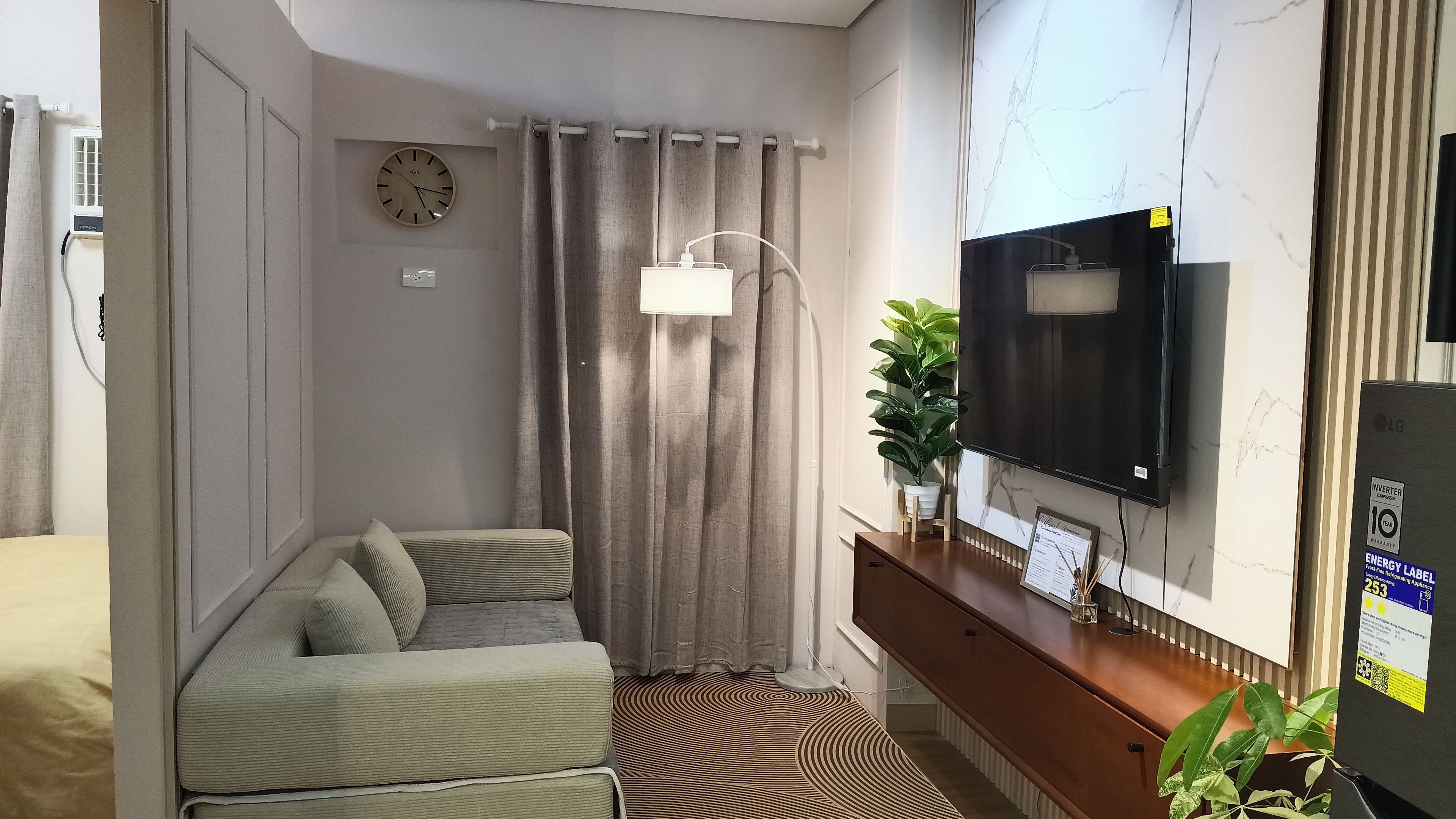 A compact living space with a sofa, a television mounted on the wall, a clock, and a potted plant.