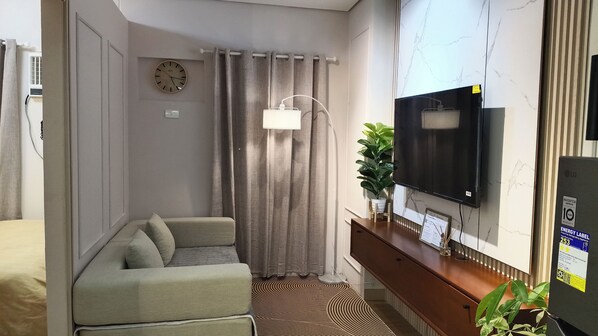 A compact living space with a sofa, a television mounted on the wall, a clock, and a potted plant. - Luxurious Casa Karina at SMDC Cheer Residences (Marilao)