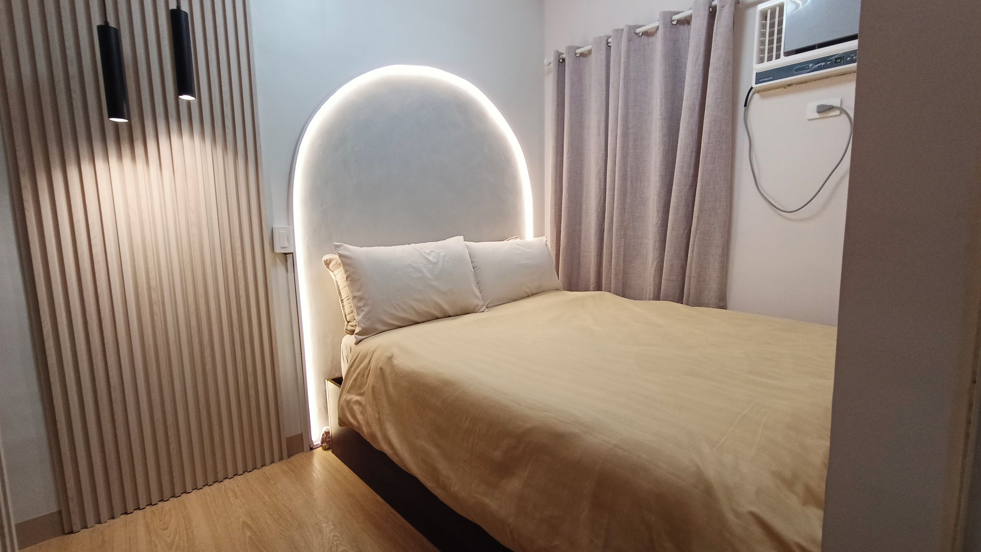 Une chambre à coucher avec un lit, une tête de lit dotée d’un éclairage circulaire, une fenêtre avec des rideaux et un climatiseur fixé au mur.