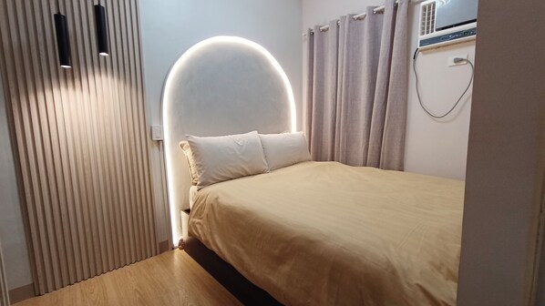 A bedroom with a bed, a headboard with a circular light, a window with curtains, and a wall-mounted air conditioner.
