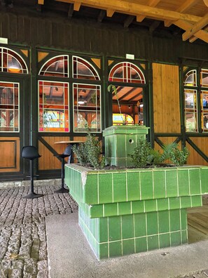 Ein grüner Pflanzkasten mit Pflanzen, eine Holzbank und ein gefliester Boden mit Steinmuster.