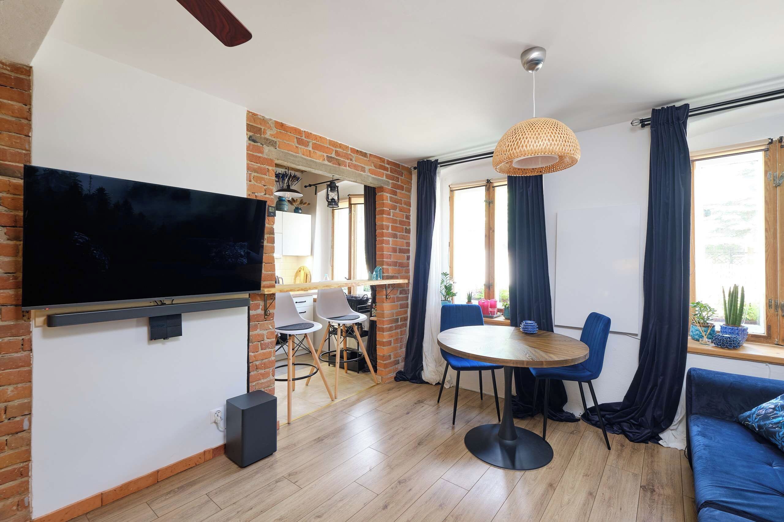 Una sala moderna con una televisión de pantalla plana, una mesa redonda de madera, sillas azules, un sofá negro y una pared de ladrillo.