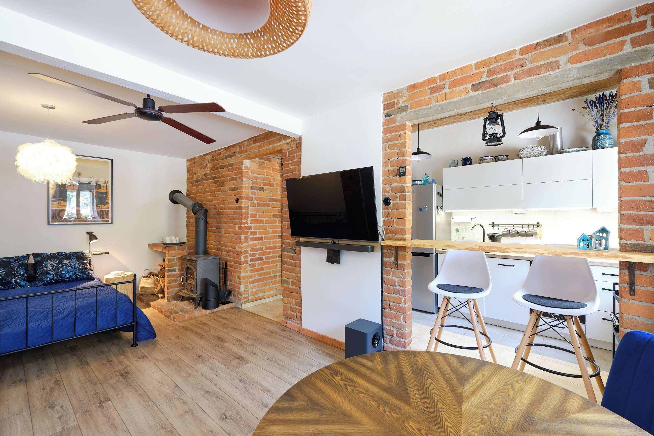 Un espacio habitable moderno con chimenea de ladrillo, un televisor de pantalla plana, una zona de comedor con una mesa redonda de madera y una cocina con armarios blancos.