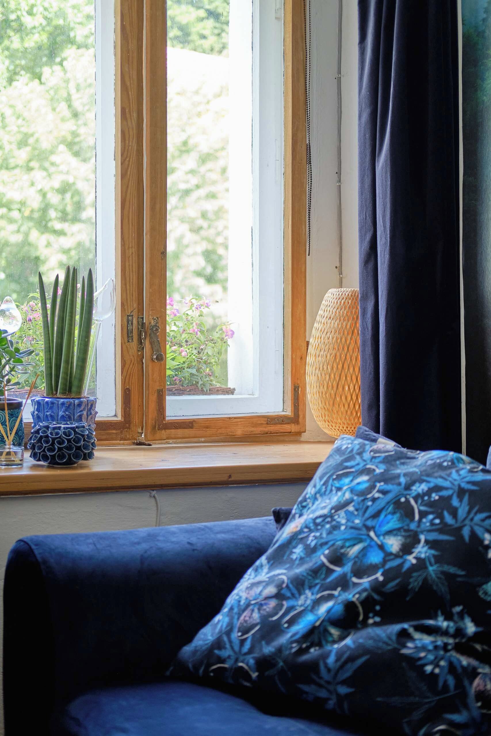 Una stanza con una finestra con cornice in legno, un divano blu con un cuscino decorato e una lampada intrecciata sul davanzale.