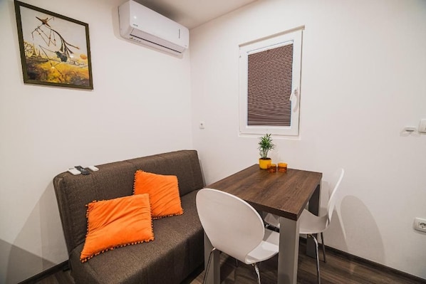 A room with a sofa, a dining table, a chair, an air conditioner, a window with blinds, and a small plant on the table.