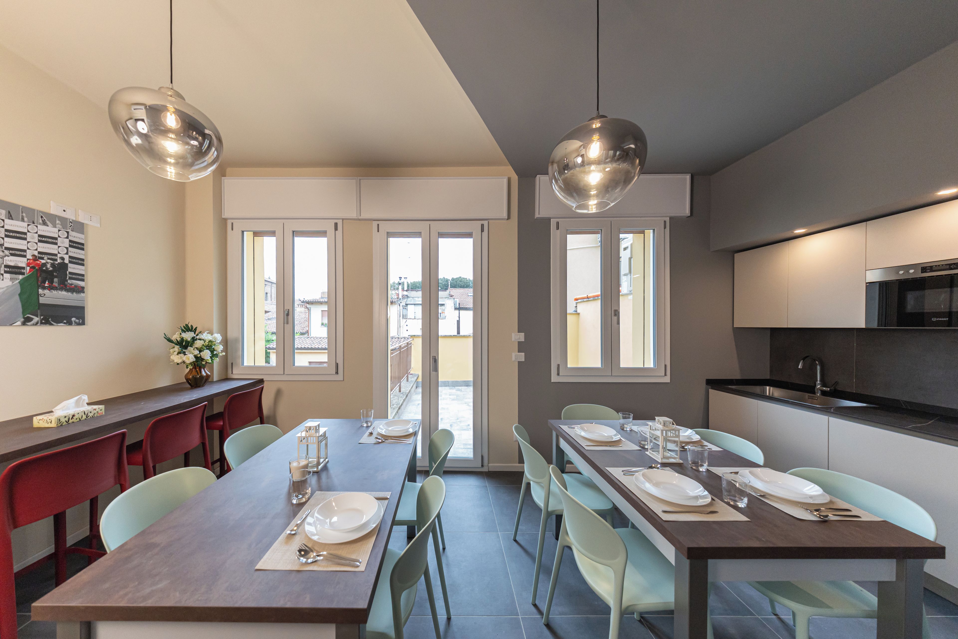 Un comedor moderno con una larga mesa dispuesta para cuatro personas, dos lámparas colgantes y vistas a través de puertas de vidrio.