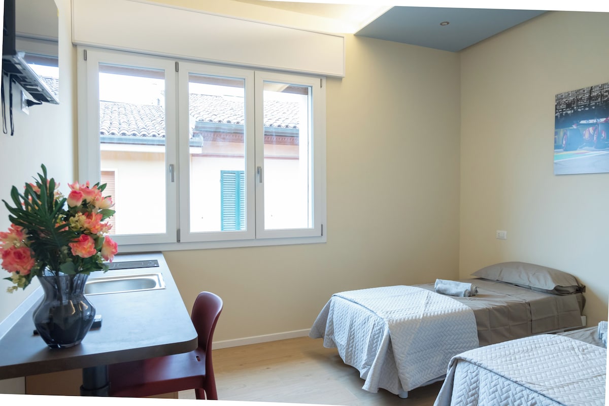 Una habitación de hotel con cama, escritorio, silla, ventana con contraventanas y un cuadro en la pared.