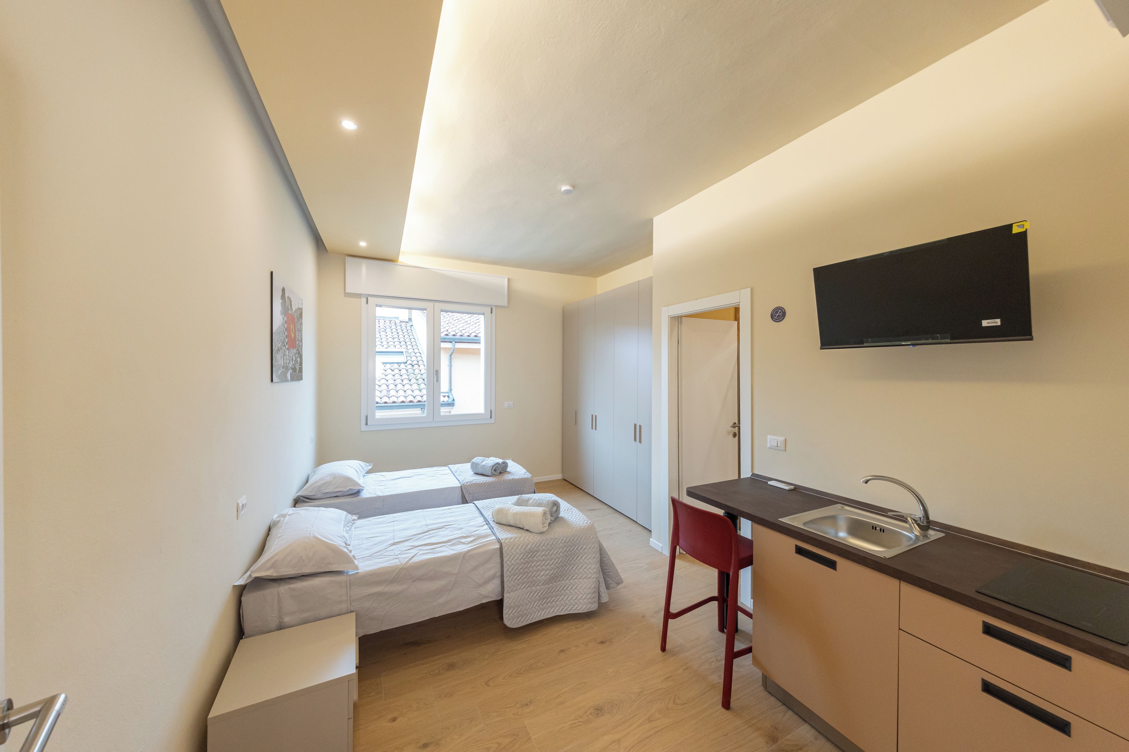 A compact bedroom with two beds, a small kitchenette, and a TV mounted on the wall.