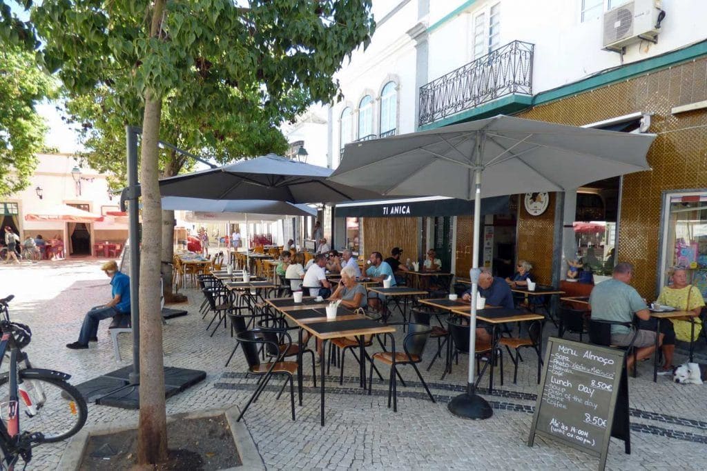 An outdoor café with tables and chairs, a menu board, and a building with a balcony in the background.