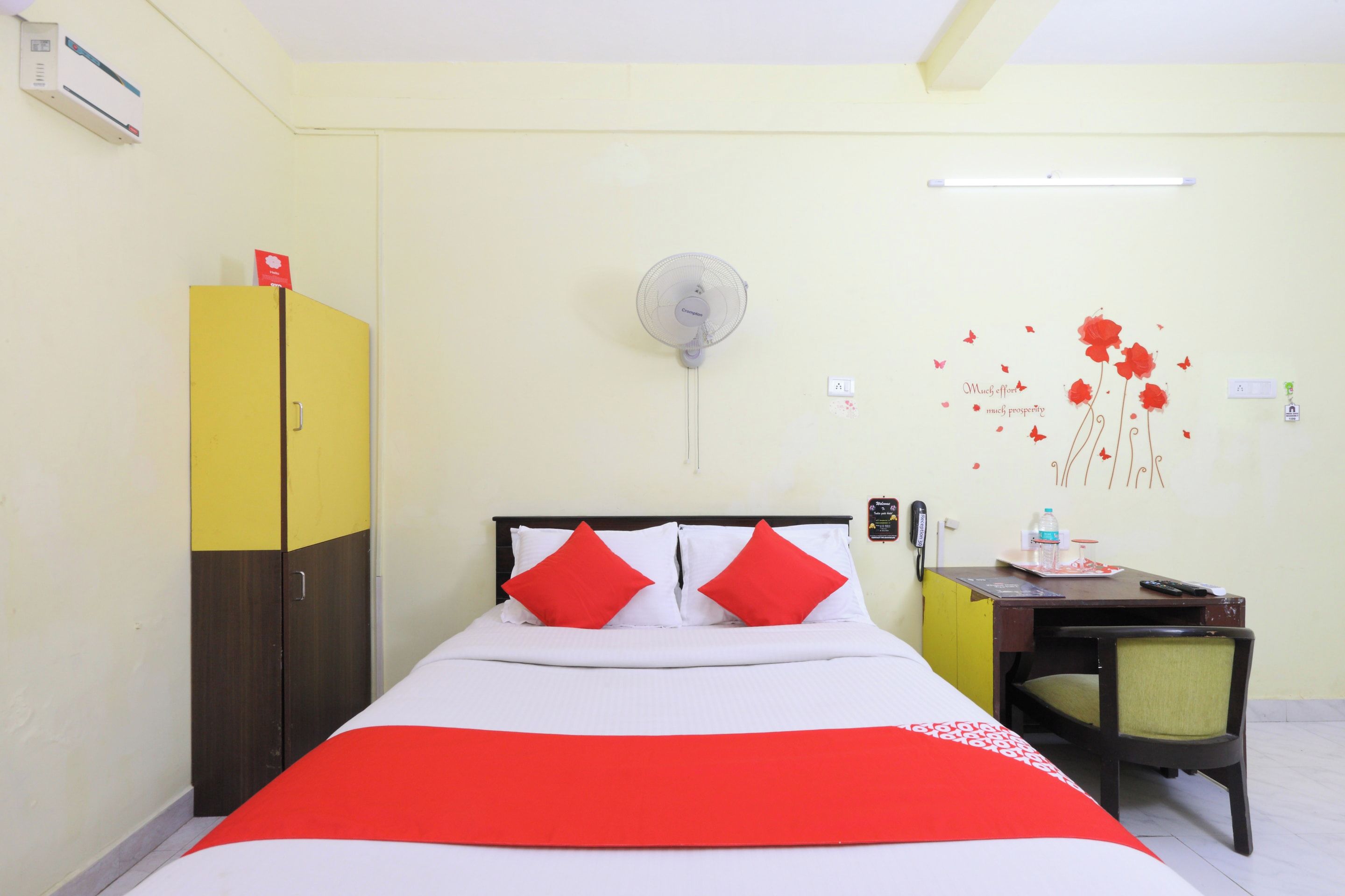 Habitación de hotel con cama, escritorio, silla, armario, ventilador de pared y un adorno en la pared.