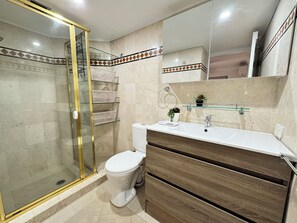 Ein Badezimmer mit einer gläsernen Duschkabine, einer weißen Toilette, einem Waschbecken mit Spiegel darüber und einer Holzkommode.