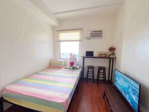 Eine kleine Einzimmerwohnung mit einem Bett, einem Schreibtisch, zwei Hocker, einem Fernseher und einem Fenster mit Jalousien.