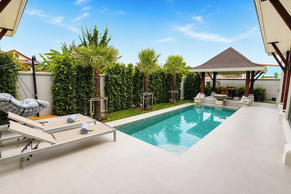 Een zwembad met ligbedden en een pergola, omgeven door groen en een heldere lucht.