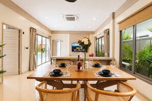 A dining area with a wooden table set for four, surrounded by wicker chairs, with a large window offering a view of greenery. - Tropical Escape New 2BR Villa with Private Pool (Tambon Rawai)