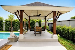 Een overdekt terras met een zwembad, houten meubels en een pergola.