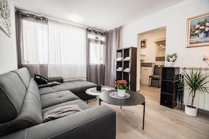 Una sala de estar moderna con un sofá gris, una mesa redonda de centro y una mesita auxiliar. Se ve una estantería con ropa doblada y una planta en maceta.