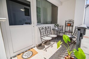 Zona exterior petita amb caires de metall, una taula, una planta en test i una finestra amb cortines.