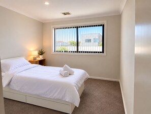 A hotel room with a bed, a nightstand with a lamp, a window with blinds, and a view of buildings outside. - Stylish 4-Bed Terrace | Modern & Convenient (Throsby)