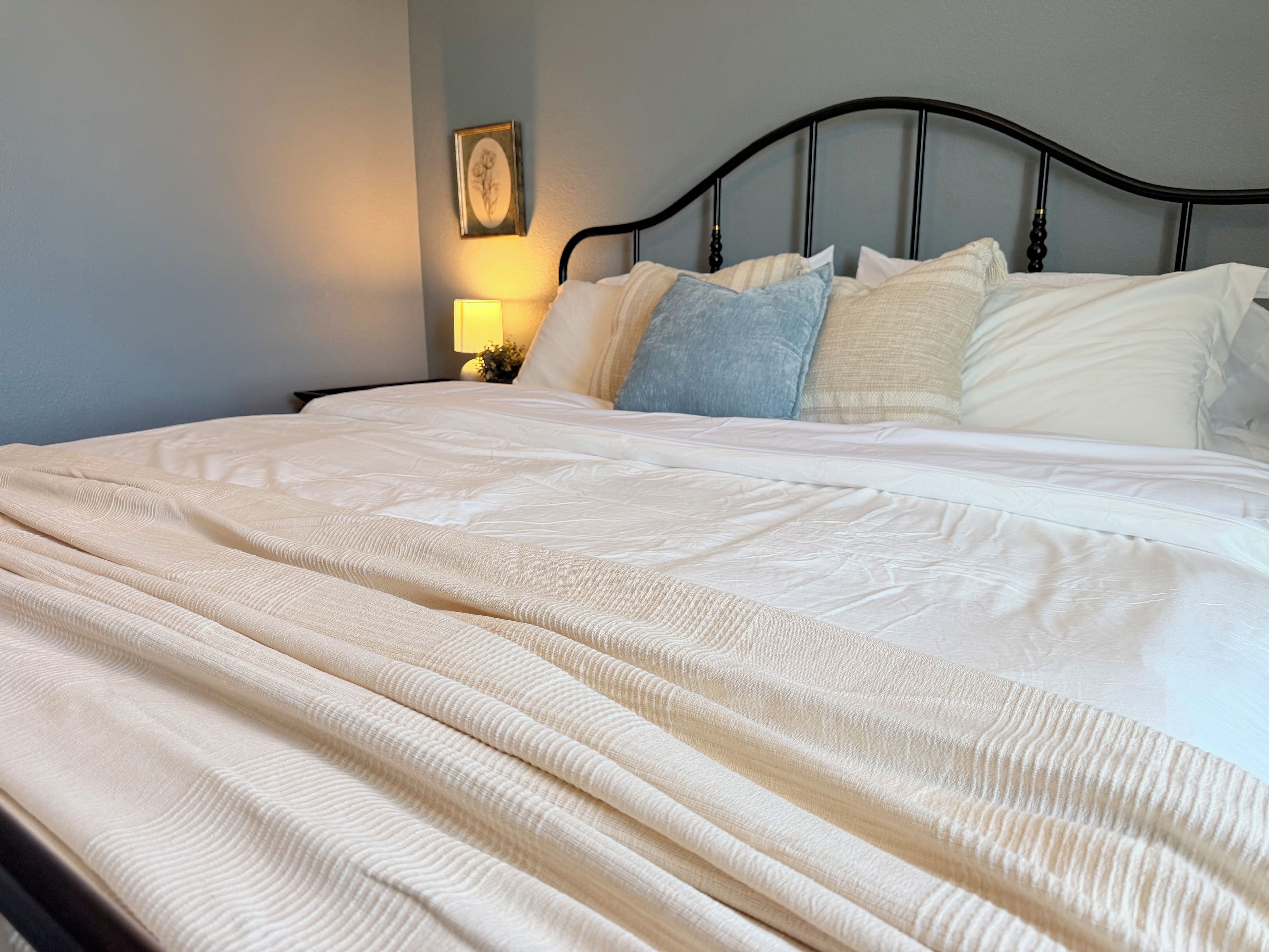 Una cama bien tendida con una colcha blanca y almohadas, un cabecero de metal negro y una lámpara fijada a la pared.