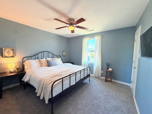 A bedroom with a bed, a nightstand, a bedside lamp, a ceiling fan, a window with curtains, and a door.