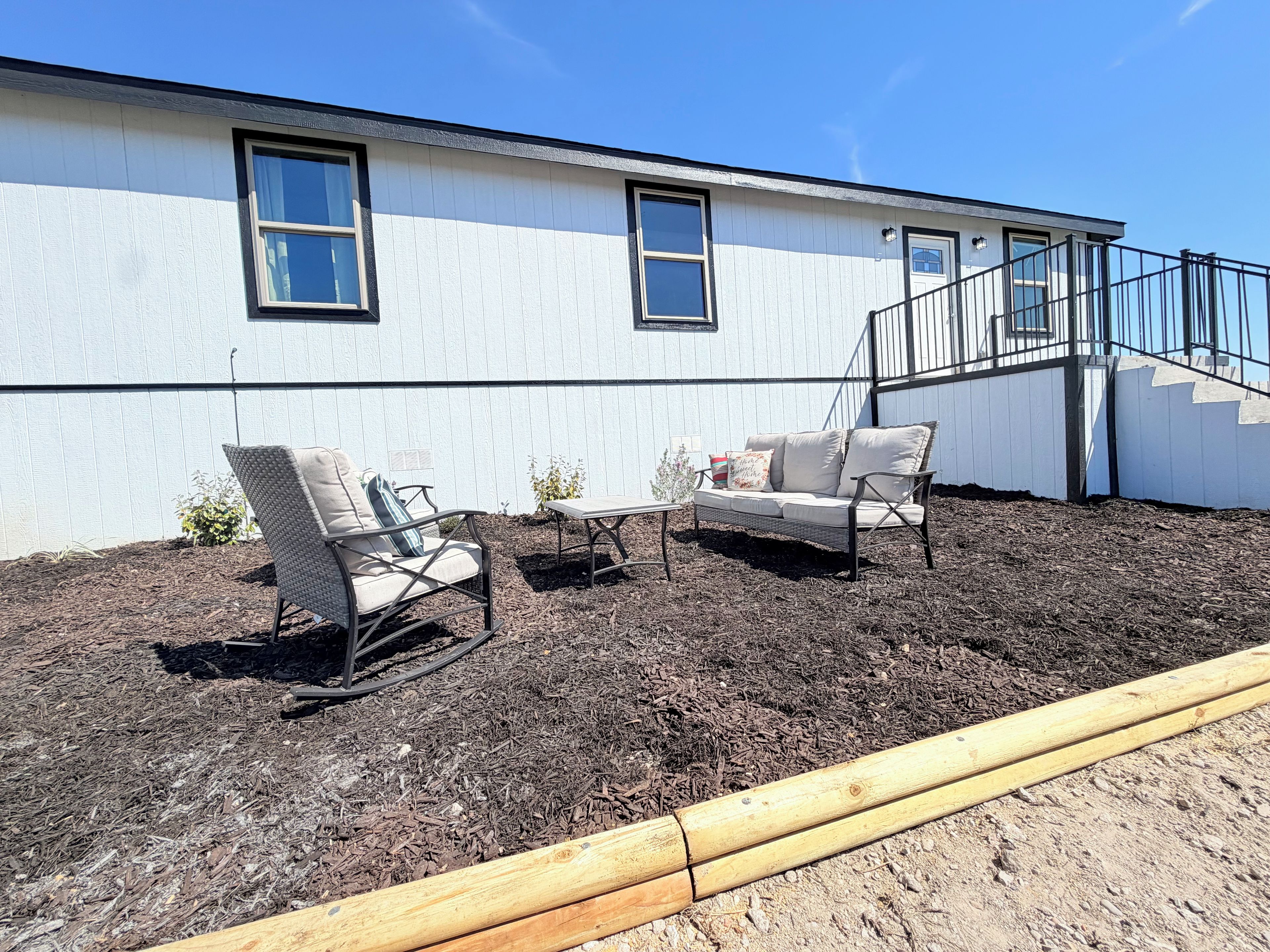 A patio area with two chairs and a table, a wooden deck, and a white building with windows and a staircase.