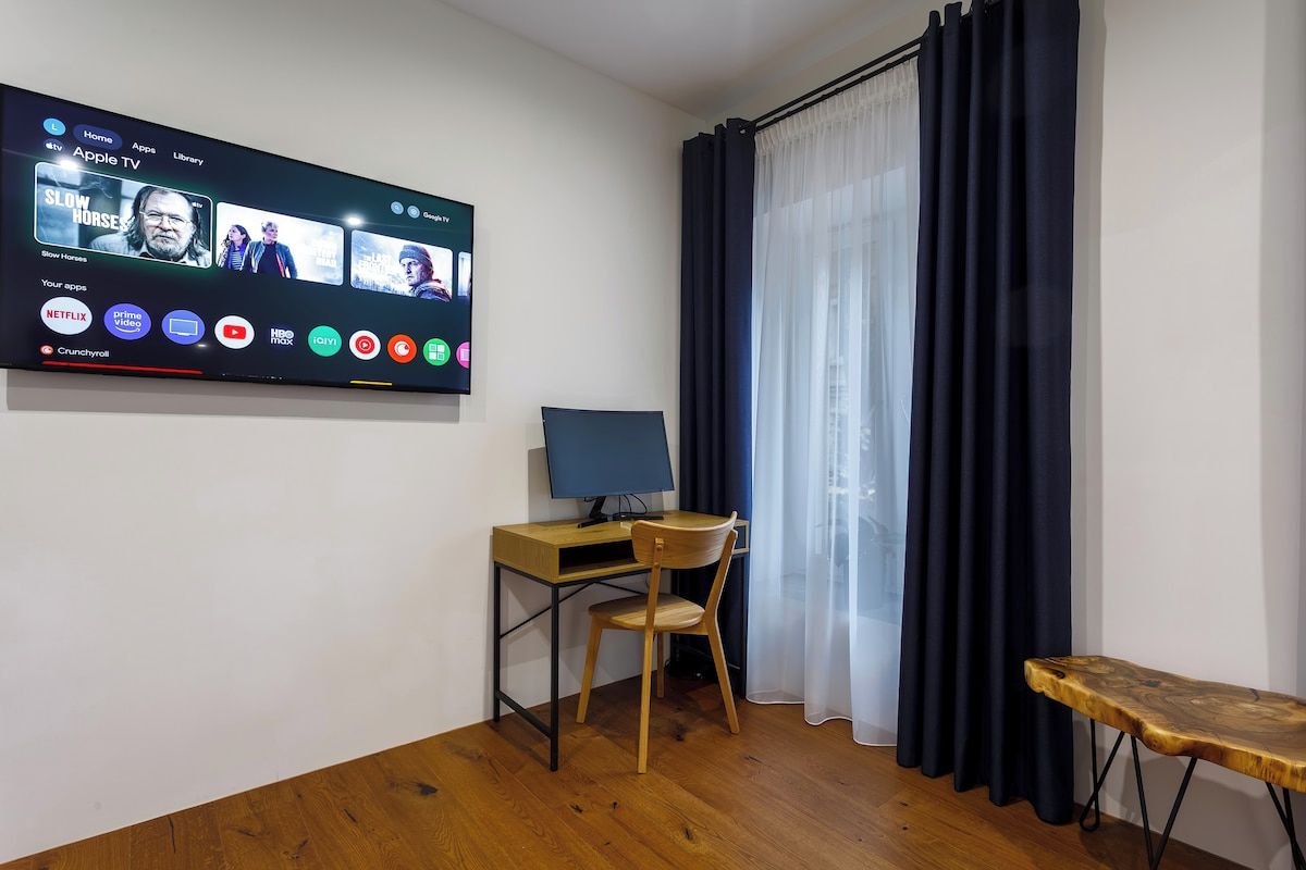 Et værelse med et fladskærms-tv på væggen, der viser forskellige apps og en film. Der er et skrivebord med en stol og en bærbar computer, et vindue med mørke gardiner og en lille træbænk.