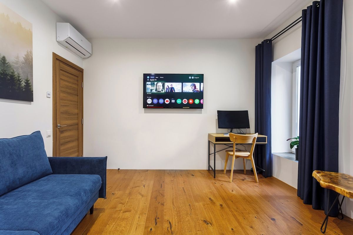 Una habitación con un sofá azul, un escritorio de madera con una computadora, un televisor montado mostrando un servicio de streaming y una ventana con cortinas.